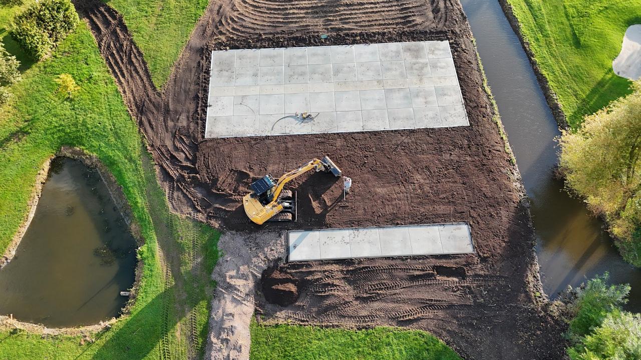 Een graafmachine is bezig met grondwerkzaamheden naast een watergang, waar grote betonnen platen al zijn geplaatst.