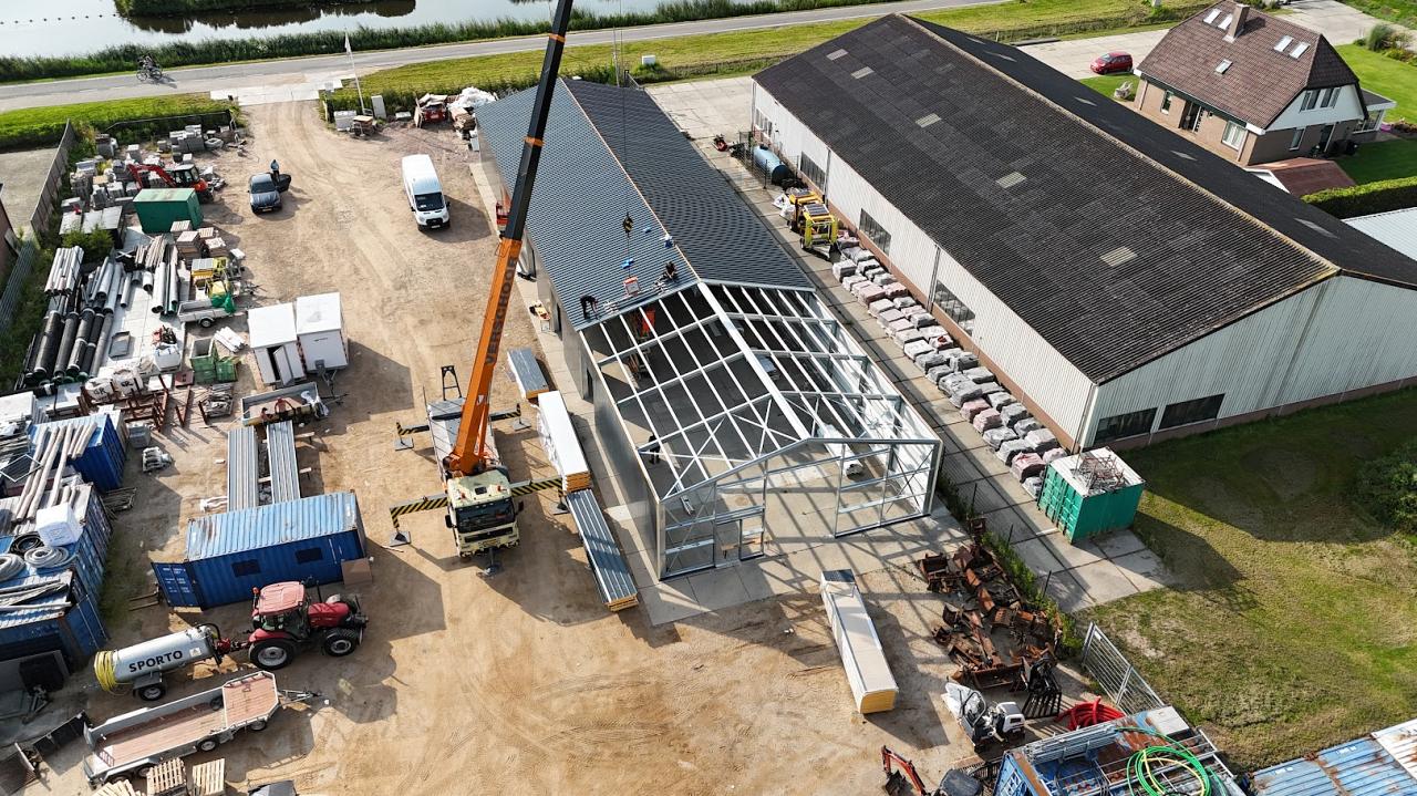 Een bouwkraan plaatst dakplaten op een nieuw stalen bedrijfsgebouw naast een bestaande loods.