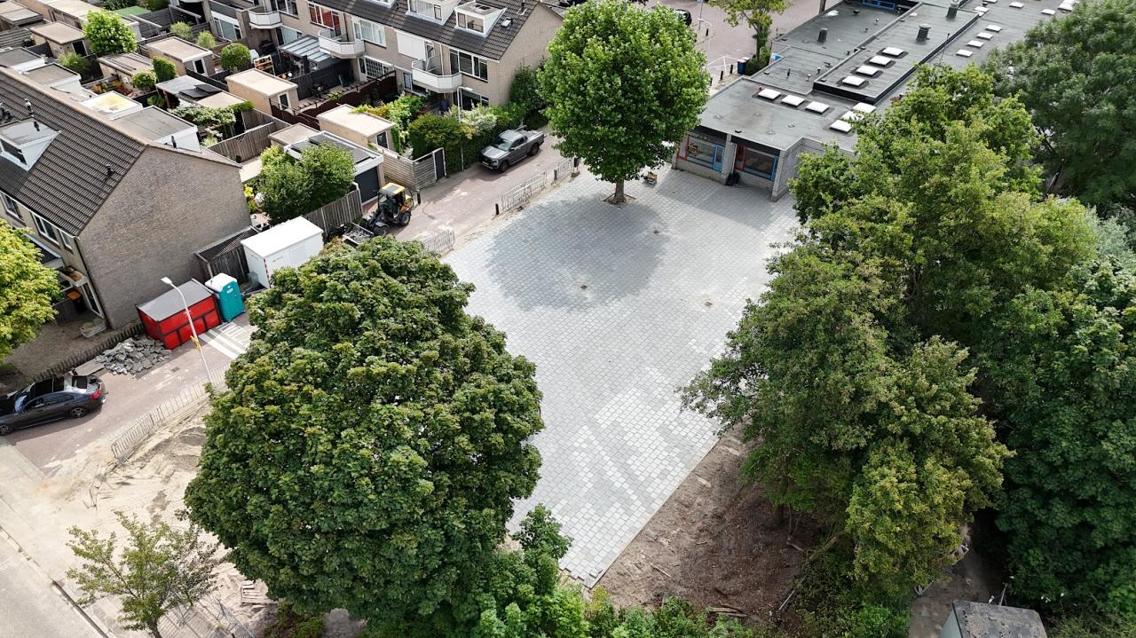 Een nieuw betegeld plein tussen woningen en een schoolgebouw, met bomen die midden in de bestrating zijn uitgespaard.