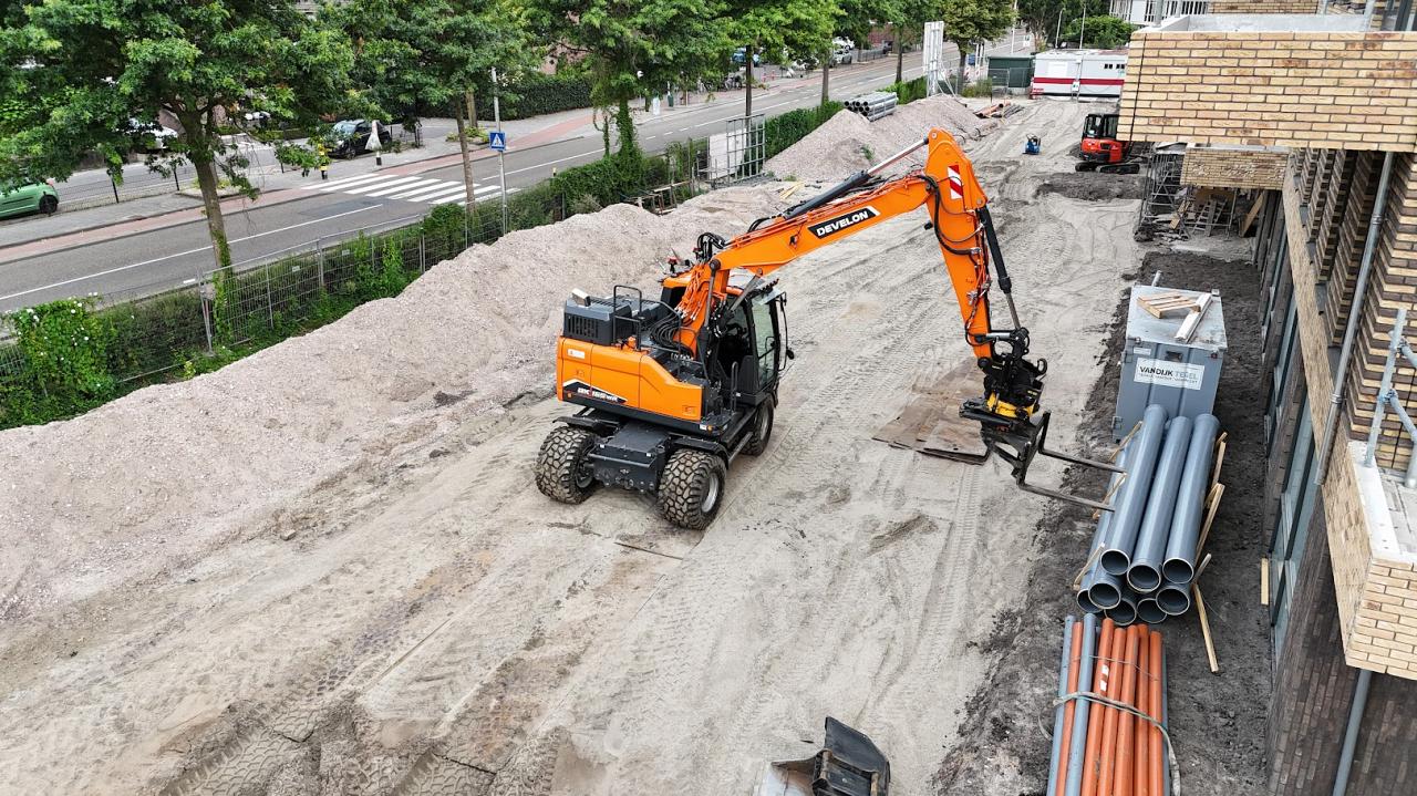 Een graafmachine plaatst grijze pvc-buizen langs een nieuwbouwlocatie naast een drukke straat.