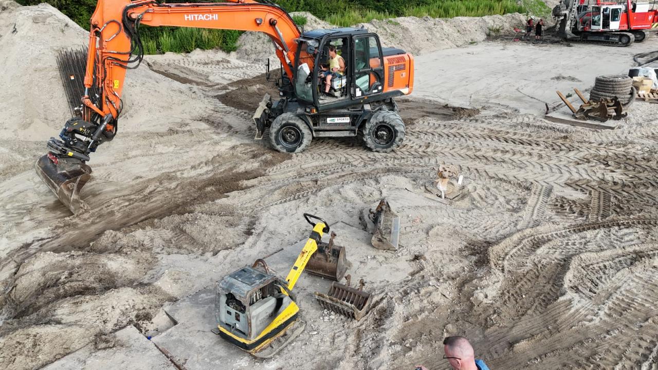 Een graafmachine van Hitachi werkt op een zandterrein, omringd door bouwmaterieel en duidelijke bandensporen in de grond.