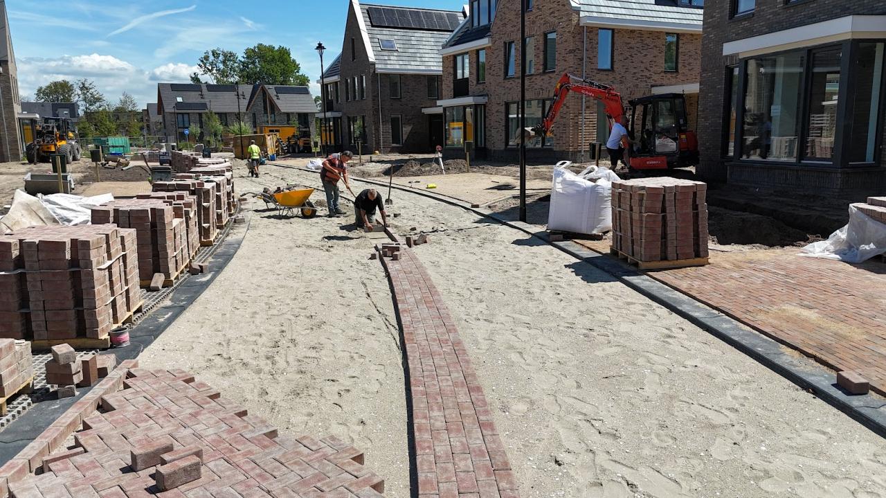 Stratenmakers leggen klinkers in een nieuwbouwwijk terwijl pallets met stenen en machines klaarstaan voor verdere aanleg.