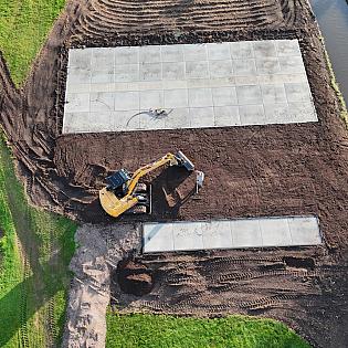 Een graafmachine is bezig met grondwerkzaamheden naast een watergang, waar grote betonnen platen al zijn geplaatst.