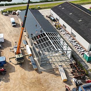 Een bouwkraan plaatst dakplaten op een nieuw stalen bedrijfsgebouw naast een bestaande loods.