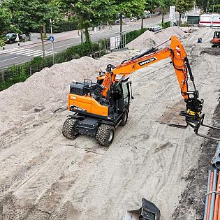 Een graafmachine plaatst grijze pvc-buizen langs een nieuwbouwlocatie naast een drukke straat.