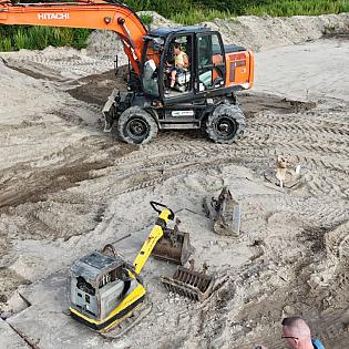 Een graafmachine van Hitachi werkt op een zandterrein, omringd door bouwmaterieel en duidelijke bandensporen in de grond.
