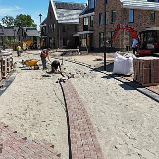 Stratenmakers leggen klinkers in een nieuwbouwwijk terwijl pallets met stenen en machines klaarstaan voor verdere aanleg.